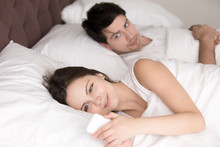 Couple In Bed, Happy Smiling Woman Turned Her Back To Man, Reading Message On Phone From Her Lover, Worried Boyfriend Lying Next To Her, Trying To Peek At Screen. Cheating And Infidelity Concept