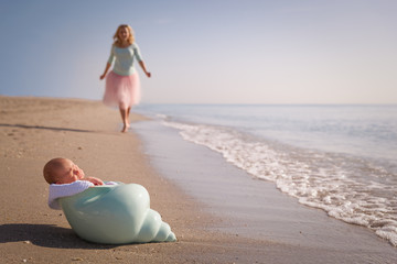 Beautiful woman running to her newborn baby boy on the sandy ocean beach. Happy mother meeting her child. Newborn baby in the nutshell on the sea shore on sunny day. Motherhood and childhood concept.