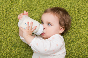 Wall Mural - baby drinking milk from bottle lying on a green carpet. Baby holding bottle himself. sweet funny baby drinking.