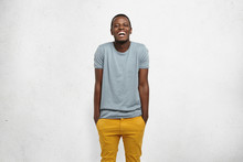 Human Feelings And Emotions. Body Language. Studio Shot Of Young Cheerful African Male, Dressed Casually, Hands In Pockets Of Yellow Pants, Shrugging, Laughing At Camera, Isolated On White Background