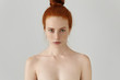 Head and shoulders of attractive young female model with ginger hair bun and freckles posing topless at blank wall. Beauty and skincare concept. Beautiful redhead girl wearing no clothes in studio