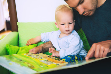 A little boy is reading a book with his father in a home interiors.