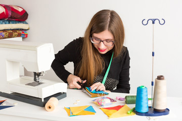 needlework and hand quilting in the workshop of a young tailor on white background - woman cuts with scissors stitched fabric at the desk with a sewing machine, spools of thread and pin cushion