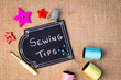 Sewing Tips on blackboard with buttons, cotton thread reels and other items on burlap background
