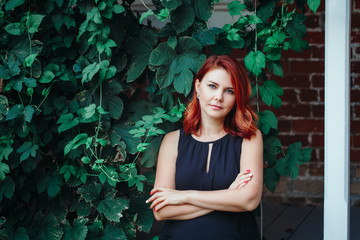 Closeup portrait of pensive middle aged white caucasian woman with waved curly red hair in black dress, looking in camera, outside in green park garden, beauty fashion lifestyle