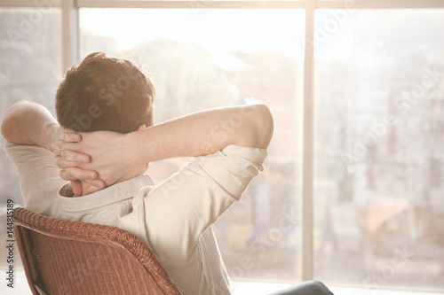 Heureux jeune homme assis sur une chaise près de la grande fenêtre Poster Mural XXL