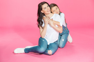 Happy beautiful mother and daughter sitting together and hugging on pink