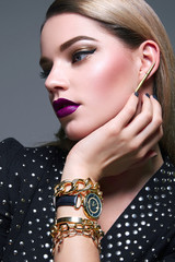 Wall Mural - Beautiful young well-groomed girlfriend with smooth hair sitting on a gray background. Dress with metal ornaments. On his hand are gold chain bracelets and watches. Evening make-up, purple lips. 