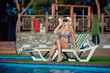 © satyrenko - Beautiful girl is sunbathing in swimsuit with pleasure. She is lying near a swimming pool. The lady is enjoying the sun and smiling. She is wearing sunglasses and a hat