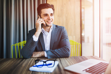 Wall Mural - Portrait of handsome young male in glasses sitting at office desk with laptop computer and talking on mobile phone.