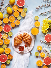 Variety of fresh citrus fruits for making juice or smoothie with fresh croissants and juice on a light blue background with spring flowers. Breakfast in the summer spring sunny mood.