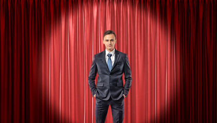 A confident businessman standing in front view in the spotlight with a red stage curtain behind him.
