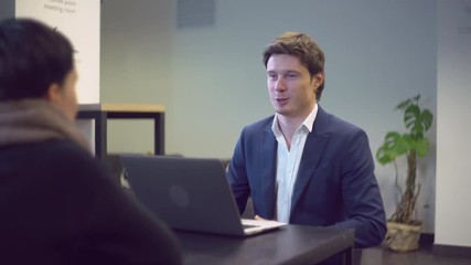 Poster - friendly interviewer asking question to woman. Handsome smiling talking with candidate for work. People sitting at the desk in modern room opposite each other.