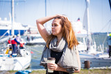 Fototapeta Młodzieżowe - Portrait of smiling stylish middle aged woman walking along sea bay. Sailboats in harbour, seaside. Sunny summer day. Wind in the hair, drinking coffee. Vacation destination. Helsinki. Finland