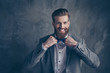 Happy young happy bearded man with mustache in formalewear stands on a gray background and holds his bow-tie with both hands
