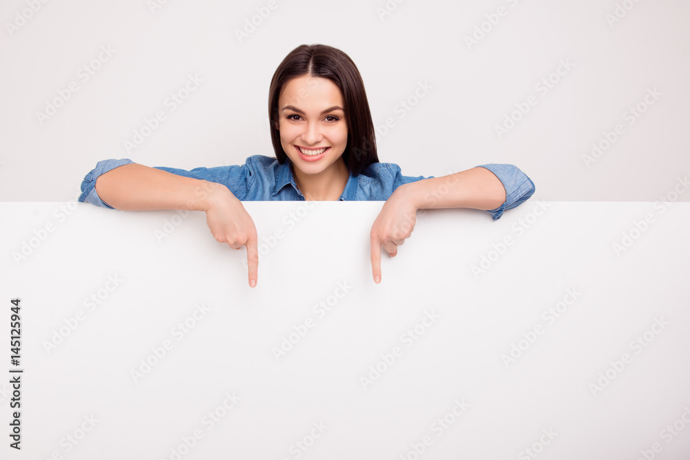 Cheerful cute girl is standing behind the white blank banner and pointing down at a copyspace - obrazy, fototapety, plakaty 
