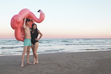 Young Couple With Inflatable Pink Flamingo On The Beach At Sunset