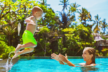 Happy child in action - active kid swim with fun in swimming pool. Baby son jump high to mother catching hands. Healthy family lifestyle, summer vacation water sports activity and lessons with parents