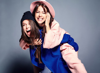 fashion portrait of two smiling brunette women models in summer casual hipster overcoat posing on gray background. Girls holding each other on back