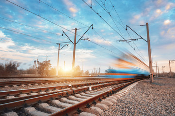 Wall Mural - High speed blue passenger train in motion on railroad at sunset. Blurred commuter train. Railway station against colorful sky. Railroad travel, railway tourism. Rural industrial landscape. Vintage