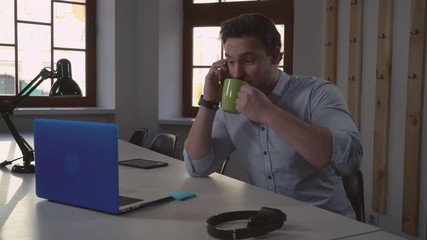 Poster - adult man using smartphone in the office. Handsome geek sitting at the desk with laptop has conversation and enjoy coffee. Businessman with beard wearing casual shirt.