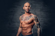 Athletic shaved head male with tattoos on his torso posing over grey vignette background.