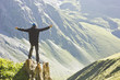 © sergeyonas - man in black jacket standing on stone with hands-up above mountais at sunset
