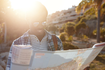 Outdoor shot of young smiling dark-skinned good-looking student on his summer journey in fashionable sunglasses and hat reading city guide standing in sun rays, isolated on blurred background