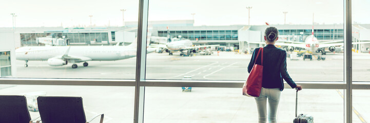 Traveler businesswoman at airport banner. Travel lifestyle. Travel tourist standing with luggage at airport lounge. Unrecognizable woman looking at window waiting at boarding gate before departure.