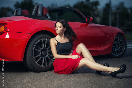 Plakat Śliczna młoda brunetka kobieta siedzi na ziemi w pobliżu koła samochodu luksusowego czerwonego kabrioletu.