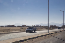 Two Old Cars Are Driving Along The Road Along The Desert And A Small Town In The Distance Against The Backdrop Of The Mountains