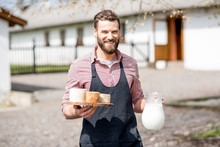 Portrait Of A Handsome Farmer In Apron Walking With Goat Cheeses And Milk Outdoors On The Rural House Background