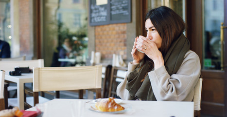 One morning a beautiful elegant woman eats breakfast at the outdoor cafe with a coffee and croissants while reading the news or emails on his tablet. Concept: Breakfast, relax, coffee, business.	