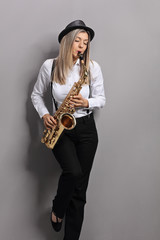 Poster - Young woman playing saxophone and leaning against gray wall