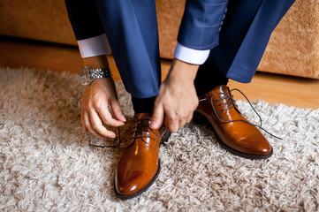 A man ties up his shoelaces on his brown shoes in the room.