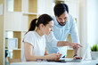 © pressmaster - Portrait of adult Asian man leaning to help young woman at desk in office and discuss work, pointing at laptop