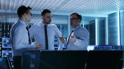 Poster - Three Technical Moderators Have Active Discussion in Monitoring Room. System Control Room is full of Working Displays Showing Various Data and Has Servers Racks. 