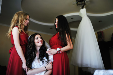 Happy cheerful brunette bride wearing at her room with two bridesmaids on red dress.