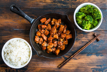 Wall Mural - Chicken teriyaki pan-fried in a cast iron skillet  with garnish of rice and broccoli in white bowls. Chopsticks, wooden rustic table, top view.