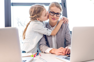 young mother and adorable daughter hugging and kissing in office with laptops