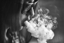 Vape. Young Handsome White Girl In Sunglasses Is Admiting Puffs Of Steam From The Electronic Cigarette. Vaping. Teenager. Black And White Photo.