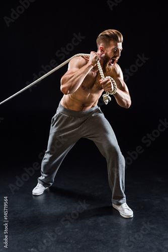 Fotografía handsome bearded sportsman yelling and pulling rope isolated on black