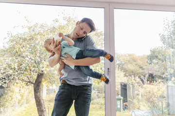 Father holding toddler son while on phone at home, by window