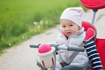 One year old baby girl sitting in a red and grey tricycle in the park, against the sunlit grass