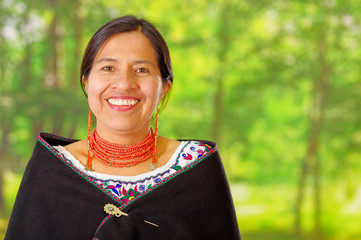 Closeup beautiful hispanic woman wearing traditional andean white blouse with colorful decoration around neck, black poncho, matching red necklace and ear, posing happily for camera, garden background