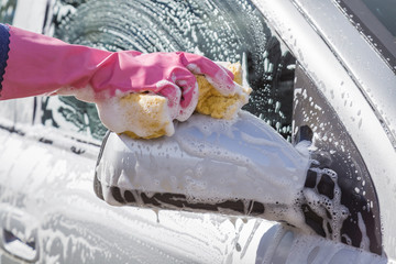 Hand in pink protective glove washing a car's mirror with sponge in sunny day. Early spring washing or regular wash up. Professional car wash by hands.