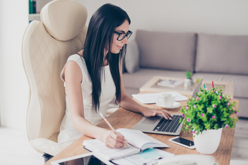 Close up portrait of smart brunette girl in glasses browsing info in laptop and writing it down in notebook at nice modern office