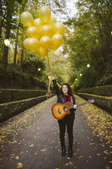 Portrait of beautiful smiling woman posing with a guitar holding yellows balloons on forest . Girl wearing black jacket, black trouser and black boots, fashion lifestyle.