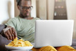 Candid shot of serious concentrated single male in hat and glasses relaxing in his apartment with laptop computer and plate of chips, surfing net or watching series. Selective focus on man's hand