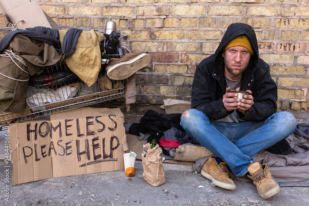 Homeless man sitting on the street, near the wall and cart with his stuff. Holding cup of coffee and looking at camera. Stock Photo | Adobe Stock